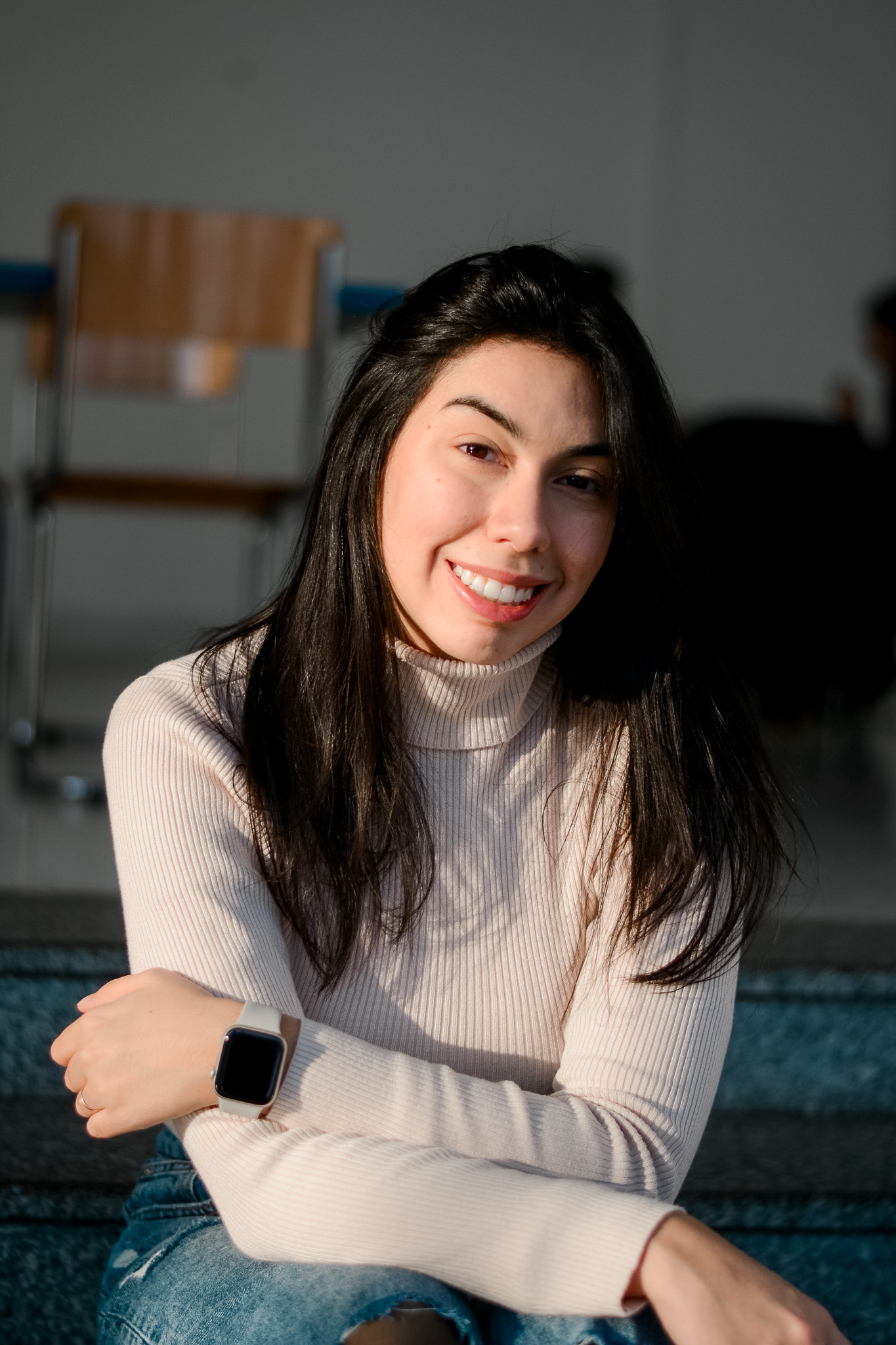 Mayara Sousa smiling at the camera, arms loosely folded, with sunlight and window shadow falling across her
