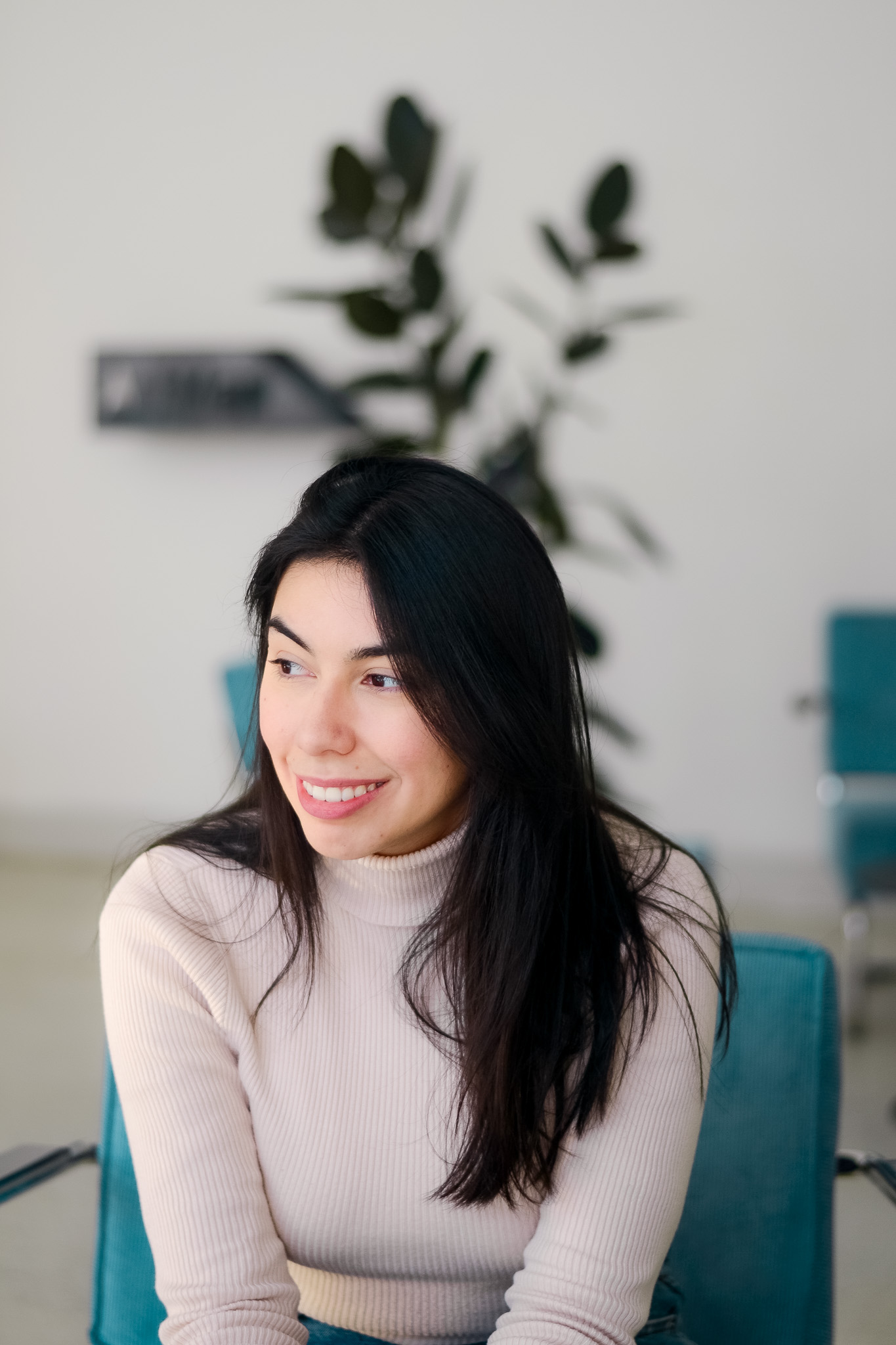 Mayara Sousa in a cream turtleneck, sitting on a teal chair in a bright studio, looking off to one side and smiling