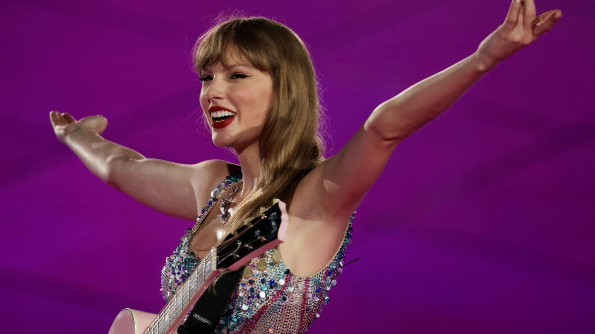 A woman with long hair smiles broadly, holding a guitar, against a vibrant pink background. She is wearing a sparkling, sequin-covered outfit and has her arms outstretched joyfully.