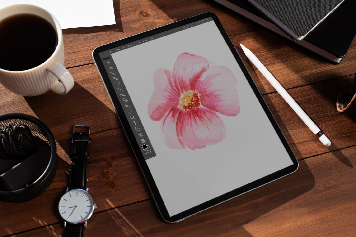 A tablet displaying a pink flower illustration sits on a wooden desk. Nearby are a stylus, a white cup filled with a dark beverage, a wristwatch, a container with paperclips, and some notebooks.